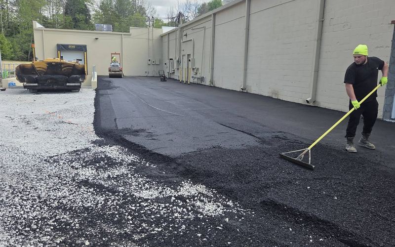 Crew member raking fresh asphalt beside a commercial building